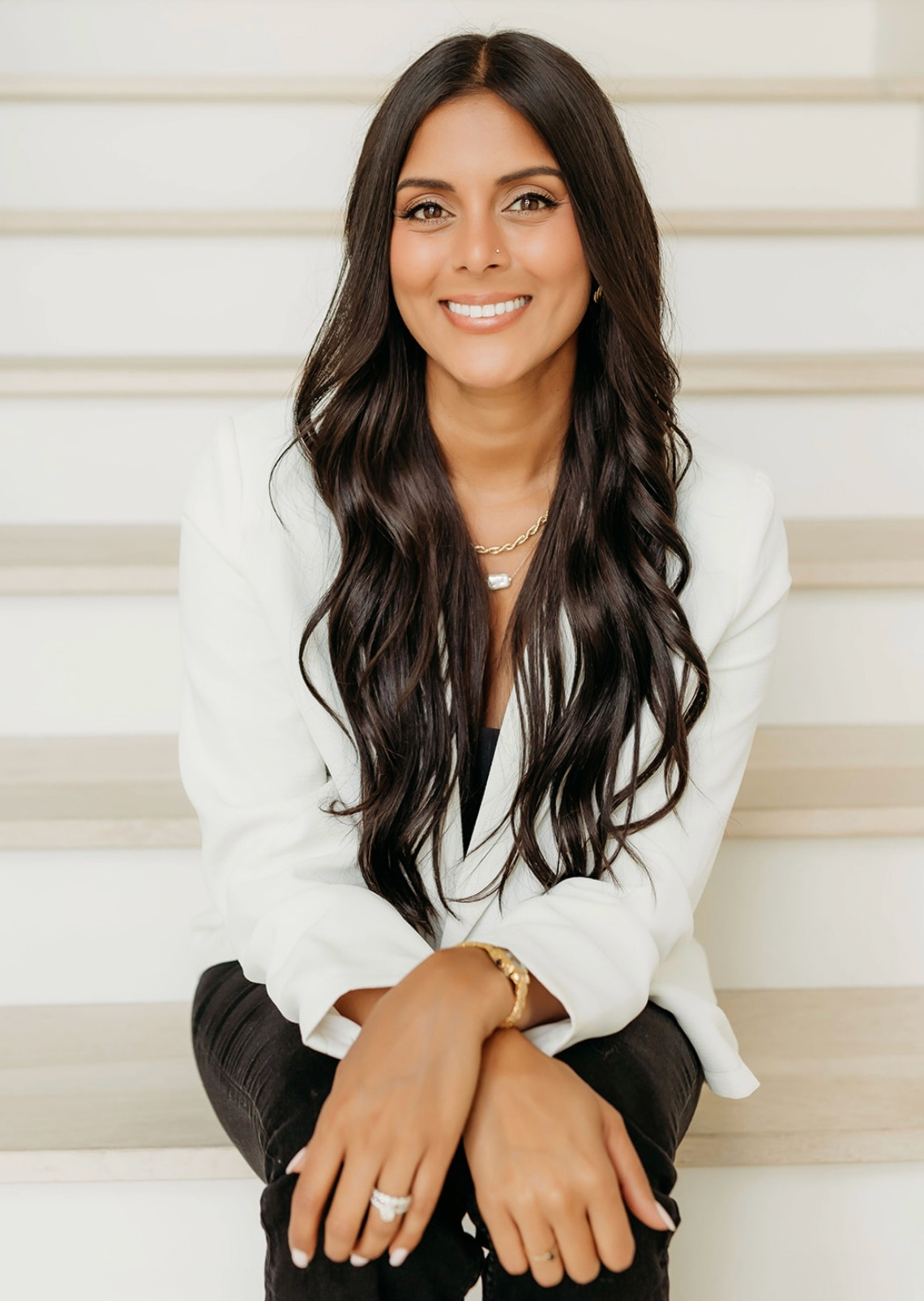 Smiling woman with long dark hair in a white blouse sitting on stairs.