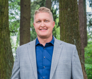 Smiling man in a gray blazer and blue shirt outdoors.